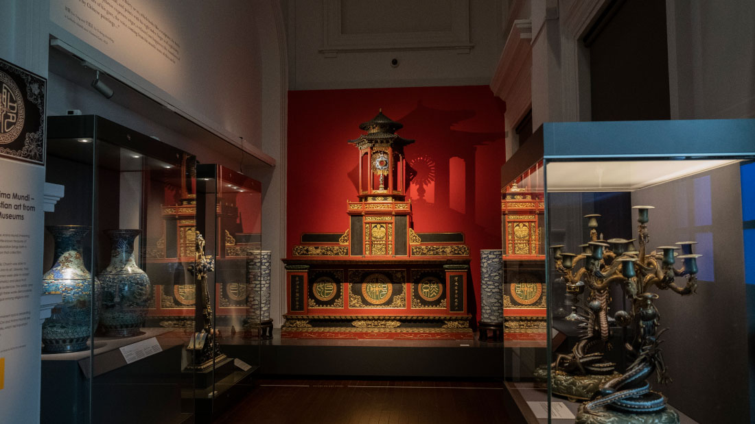 A photograph of a dimly-lit gallery space with two glass exhibition casements on the left along the left wall. One of the casement holds two large decorated vases. Another casement shows a large stylised wooden cross. In the centre of the image, there is a large red and black altar in front of a red wall. On the right side of the image, there is one glass-encased exhibit with two large candleholders.