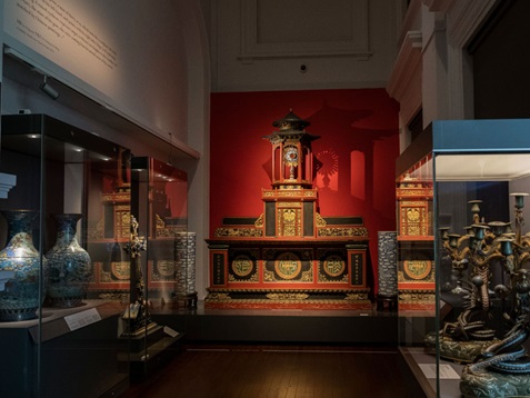 A photograph of a dimly-lit gallery space with two glass exhibition casements on the left along the left wall. One of the casement holds two large decorated vases. Another casement shows a large stylised wooden cross. In the centre of the image, there is a large red and black altar in front of a red wall. On the right side of the image, there is one glass-encased exhibit with two large candleholders.