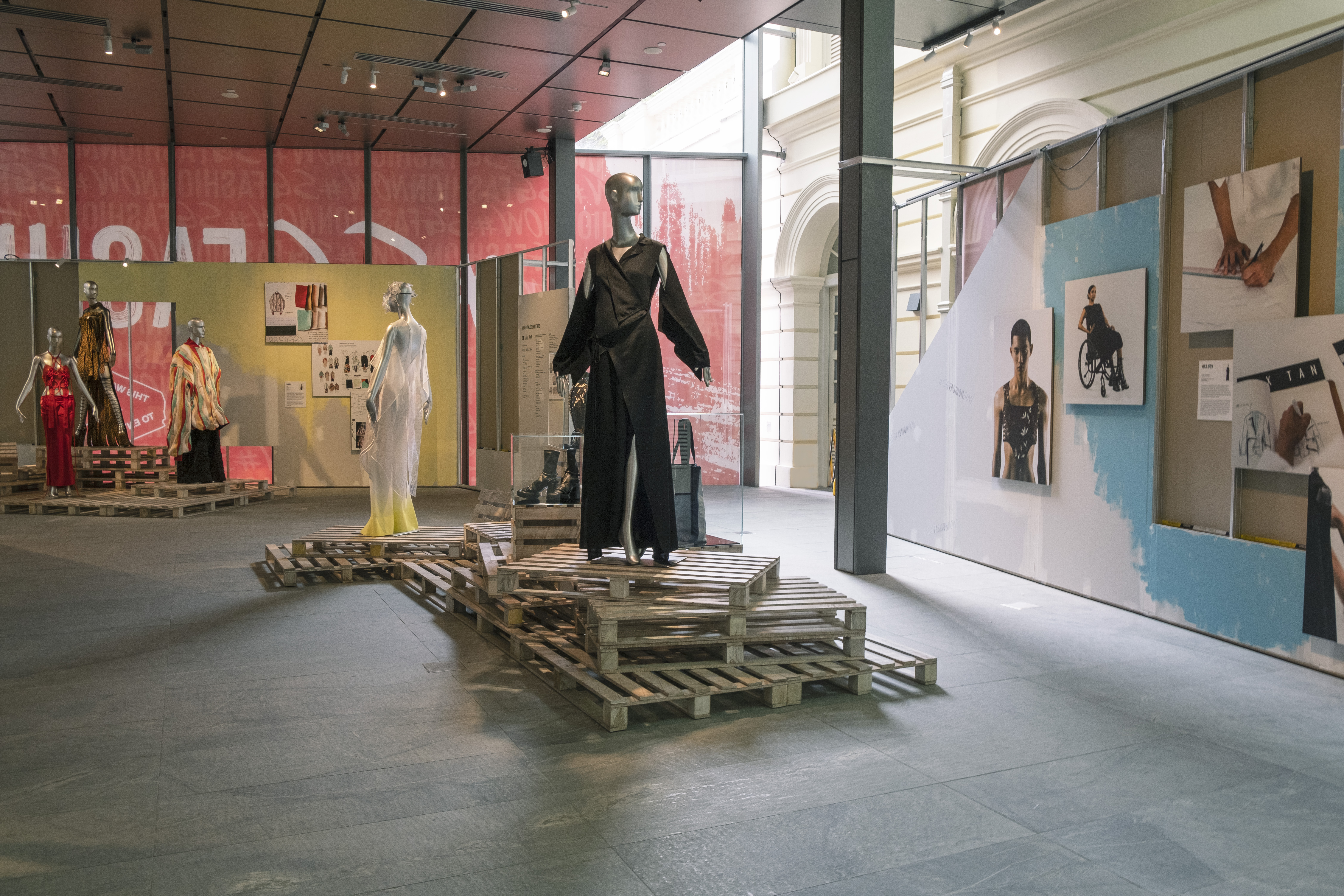A gallery space with wooden platforms on the ground and a mannequin wearing a black ensemble standing on it. The walls has posters showing the designing process. 