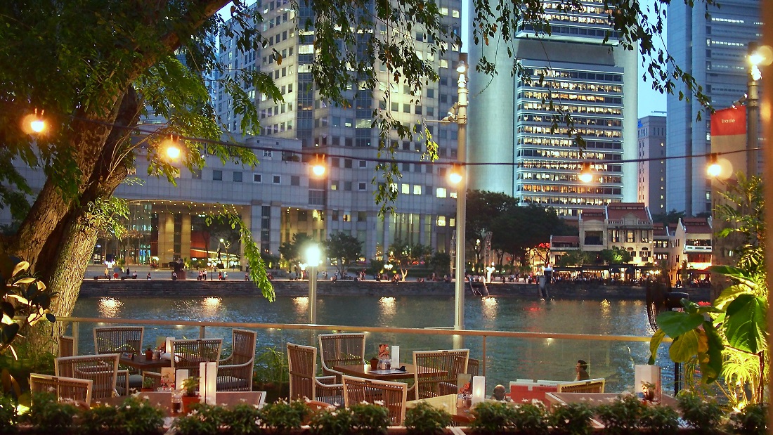 Tables and chairs outdoors by the river in the evening, with a view of skyscrapers on the other side of the river. Fairylights illuminate the area.