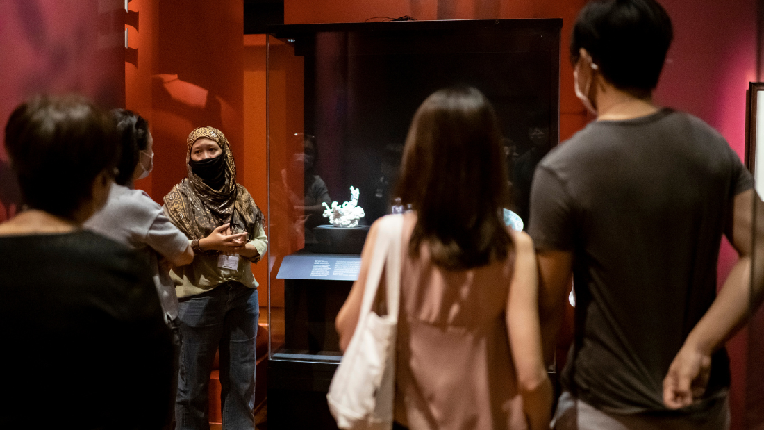 A museum docent standing in front of a glass display case in a gallery with red walls, speaking to a group of museum visitors