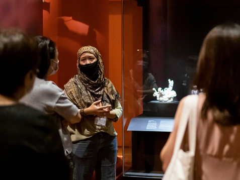 A museum docent wearing a mask in front of a display case, speaking to a group of museum visitors