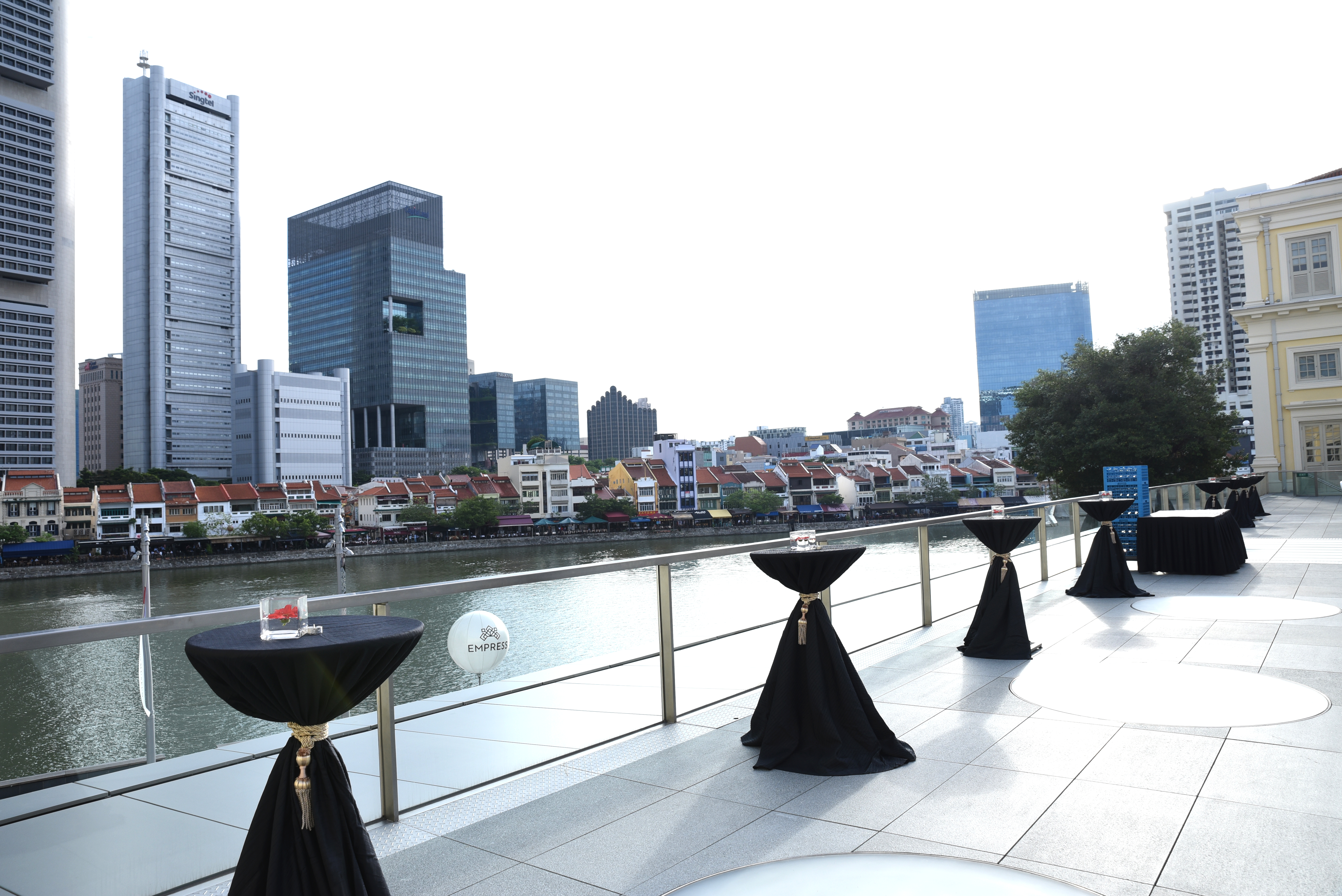 Image of an outdoor terrace with cocktail tables lined across it, and facing the Singapore River with towering skyscrapers behind 